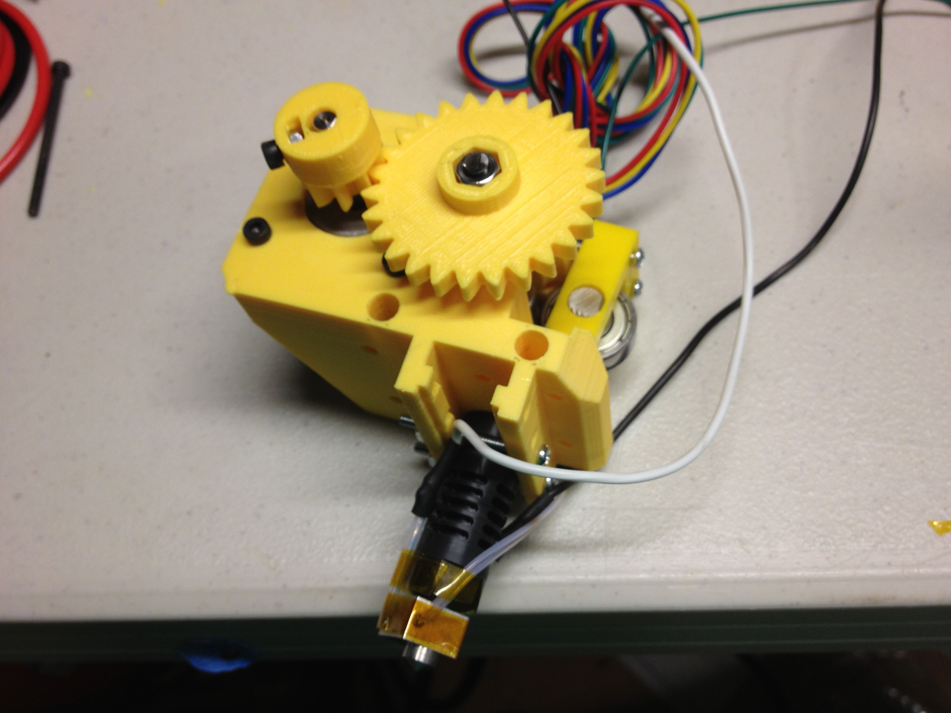 Figure 4 — An i3-lineage direct extruder on the bench: a geared drive (here the earlier single-gear style) grips the filament directly above the hotend, which hangs immediately below and feeds the …